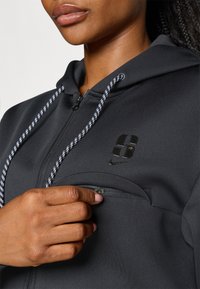 Sudadera negra con bolsillo con cremallera, logo reflectante y cordón en contraste. La tela suave y el diseño ajustado mejoran la funcionalidad.