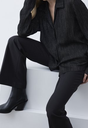 Donna che indossa una camicia nera a righe sottili, pantaloni neri e stivaletti neri con tacco, seduta su un blocco bianco contro uno sfondo bianco.