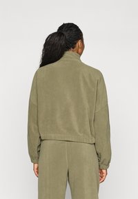 Chaqueta de forro polar verde oliva con cuello alto, hombros caídos y puños elásticos. Textura suave y longitud corta diseñada para la comodidad.