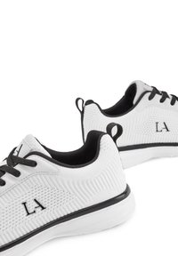 Weiße Sportschuhe mit einem strukturierten, atmungsaktiven Mesh-Obermaterial, schwarzen Akzenten und flachen Schnürsenkeln; verfügen über ein Logo an der Seite und eine Zuglasche.