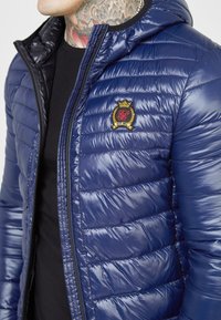 Marinblå puffjacka med en glänsande yta, med horisontellt quiltade segment, dragkedja framtill och en broderad logotyp på bröstet.