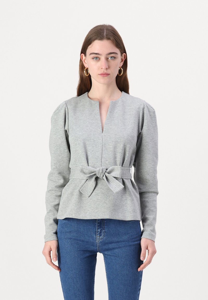 Šedý dlhý rukáv top s výstrihom do V a peplum pásom, ktorý má opasok na viazanie. Hladká textúra s jemným horizontálnym vzorom na opasku.