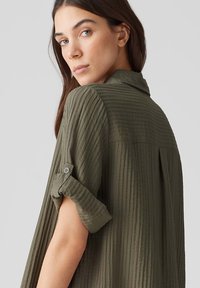 Camicia leggera di un verde oliva, testurizzata, con maniche arrotolabili, dettagli con bottoni e una silhouette rilassata. Motivo a righe verticali.