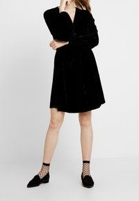Vestido de terciopelo negro con mangas largas, escote en V y cintura ajustada. Combinado con medias negras estilo red y zapatos de piso.