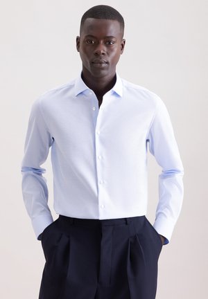 Homme portant une chemise bleu clair rentrée dans un pantalon noir plissé, debout avec les mains dans les poches devant un fond uni.