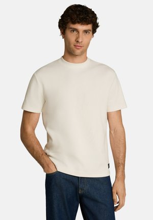 Mann mit lockigem Haar, der ein schlichtes cremeweißes kurzärmeliges Hemd und dunkelblaue Jeans trägt, steht mit einer Hand in der Tasche vor einem weißen Hintergrund.