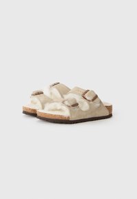 Beige suedepumper med to justerbare remmer med spenner, hvit fluffy fôr, korkfotbed og mørk gummisole.
