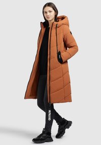 Lång, vadderad pufferjacka i bränd orange, med huva, dragkedja fram och svarta accenter. Matchad med svarta leggings och skor.