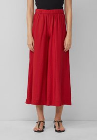 Pantalons larges rouges en tissu texturé, avec une taille élastique et une coupe ample, associés à des sandales plates noires.