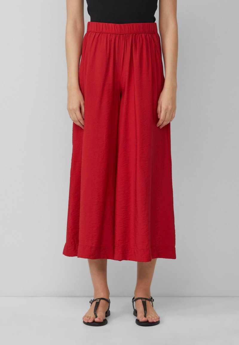 Pantalons larges rouges en tissu texturé, avec une taille élastique et une coupe ample, associés à des sandales plates noires.