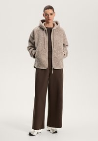 Giacca in fleece marrone chiaro con cappuccio, zip frontale e tasche laterali. Abbinata a una maglietta marrone e pantaloni larghi marroni. Le sneakers bianche completano il look.