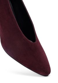 Zapato de tacón alto de ante burdeos con puntera puntiaguda y escote bajo. La textura suave y la silueta esbelta realzan el diseño elegante.