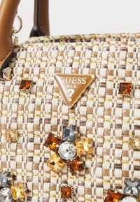 Woven handbag in beige and tan, featuring golden thread accents and multicolored gemstone embellishments; triangular "GUESS" logo attached.
