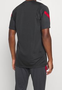 Koszulka sportowa z krótkim rękawem w ciemnoszarym kolorze z fakturowanymi czarnymi panelami na ramionach i czerwonymi akcentami na rękawach. Gładka tkanina o luźnym kroju.