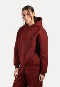 Bordová mikina s kapucí vyrobená z měkké látky, s přední kapsou a žebrovanými manžetami. V kombinaci s odpovídajícími kalhotami, které mají logo jako akcent.