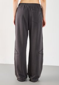 Person wearing loose, dark gray sweatpants with an elastic waist and a back pocket, visible from the waist down, standing on a plain floor.