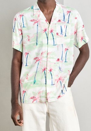 Mann trägt ein kurzärmeliges Hemd mit Pastellgrün- und Rosa-Palmenblattmuster, kombiniert mit cremeweißer Hose, steht mit einer Hand in der Tasche.