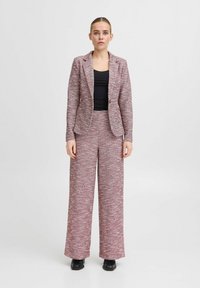 Rot-weiß gemusterter gewebter Anzug mit tailliertem Blazer und weit geschnittenen Hosen, kombiniert mit einem schwarzen Tanktop und schwarzem Schuhwerk.