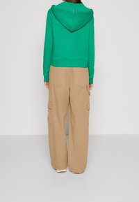 Felpa verde con cappuccio, polsini e orlo a coste, abbinata a pantaloni cargo beige con tasche laterali e vestibilità ampia.