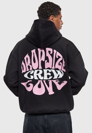 Mann trägt schwarzen Kapuzenpullover mit großem pink-weißem Schriftzug "Dropsize Crew Gove" auf dem Rücken, steht vor einem einfarbigen hellen Hintergrund.