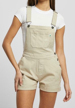 Kvinde iført beige korte overalls med frontlomme og knapdetaljer, kombineret med en hvid kortærmet t-shirt.