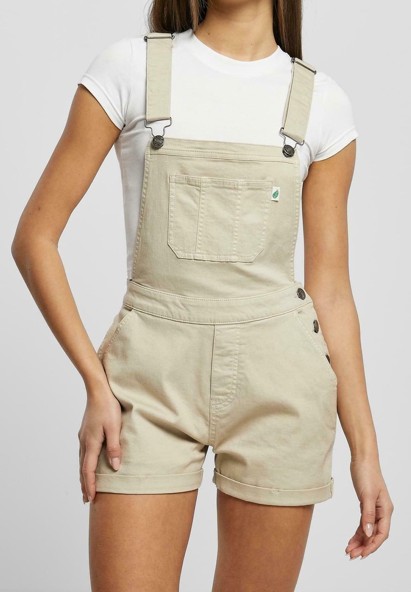 Femme portant une salopette courte beige avec poche avant et détails de boutons, associée à un t-shirt blanc à manches courtes.
