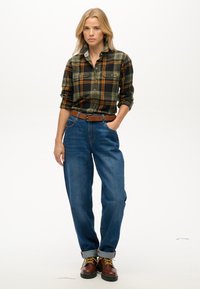 Camisa de cuadros en verde, negro y naranja; jeans azules con puños enrollados; botas marrones con cordones y acentos amarillos; cinturón de cuero marrón.