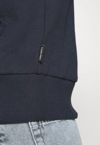 Sudadera de algodón azul marino con dobladillo y puños acanalados. Presenta una etiqueta visible de Calvin Klein en negro. Combinada con unos jeans de mezclilla claros.
