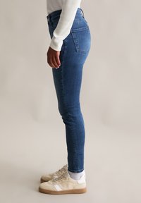 Chude niebieskie dżinsy z wysokim stanem i dwiema tylnymi kieszeniami, zestawione z beżowymi trampkami i białymi skarpetkami. Gładka faktura, dopasowany krój.