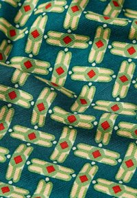 Textil con fondo teal que presenta un patrón geométrico repetido de formas verdes con acentos en diamante rojos y detalles en crema.