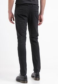 Svarta slim-fit byxor med en slät textur, med två bakfickor. Bärs med svarta dressade skor, vilket visar en ren, modern design.