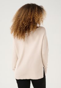 Maglione a coste di un beige chiaro con un taglio morbido, fessure laterali e maniche lunghe, realizzato in tessuto morbido, indossato sopra pantaloni neri.