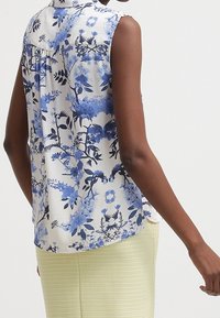 Blusa floreale con motivi blu e neri su sfondo bianco. Design senza maniche con un sottile dettaglio arricciato sul retro.