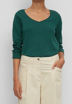 Kobieta ubrana w ciemnozieloną bluzkę z długim rękawem i dekoltem w kształcie litery V, włożoną do kremowych, sztruksowych spodni z wysokim stanem, z jedną ręką w kieszeni, stojąca na tle szarego tła.