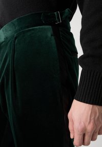 Velvet green trousers feature a high waistband with a buckle and a black side stripe. The texture is soft, adding depth to the design.