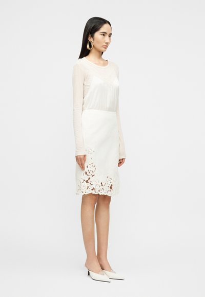 Γυναίκα όρθια που φοράει λευκή διαφανή μπλούζα με μακριά μανίκια, λευκή φούστα με κορδόνι με λουλούδια στην άκρη, μεγάλα χρυσά σκουλαρίκια και μυτερές λευκές γόβες.