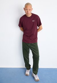 Homem careca está de pé com as mãos atrás das costas, vestido com uma t-shirt Nike bordeaux, calças de veludo verde e ténis brancos sobre um tapete azul.