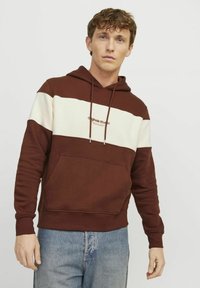 Bluza z kapturem w ciemnym brązie i kremowym, z przednią kieszenią, sznurkami do regulacji i haftowanym logo na piersi. Miękka, teksturowana tkanina.
