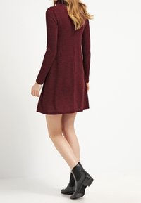 Abito lavorato a maglia bordeaux con maniche lunghe e gonna svasata; presenta una texture a costine e un leggero dettaglio a collo alto, abbinato a stivali neri alla caviglia.