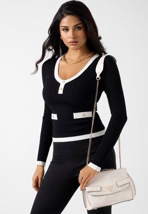 Vrouw draagt een zwarte geribbelde trui met witte bies en gouden knopen, zwarte broek, gouden ketting, en houdt een witte handtas met patroon en kettingriem vast.