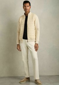 Jachetă capitonată bej deschis cu guler înalt, cămașă neagră și pantaloni de culoare crem. Pantofi slip-on din suede completează ținuta.