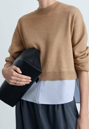 Personne portant un pull marron par-dessus une chemise blanche et un pantalon sombre, tenant une grande pochette noire avec une bague argentée à la main gauche.