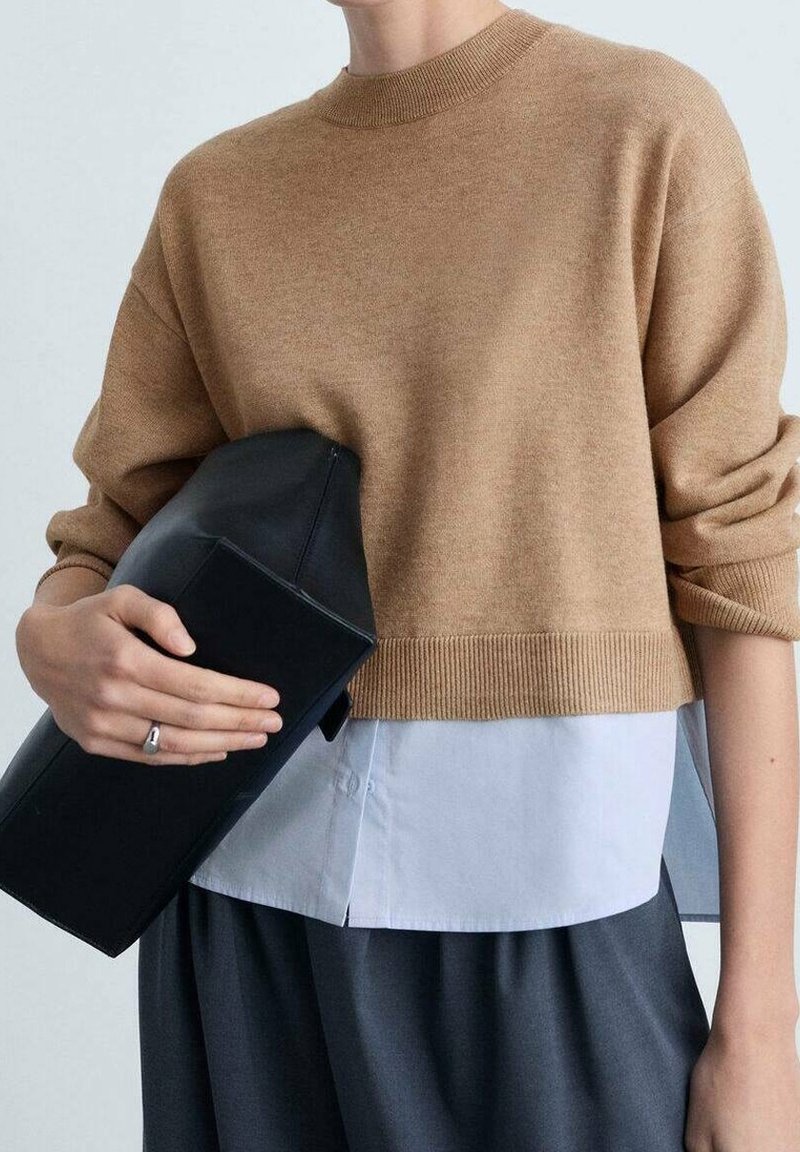 Personne portant un pull marron par-dessus une chemise blanche et un pantalon sombre, tenant une grande pochette noire avec une bague argentée à la main gauche.