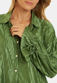La blusa verde plisada cuenta con un cuello tipo chal, una tela texturizada y mangas abullonadas con detalles fruncidos y botones dorados.