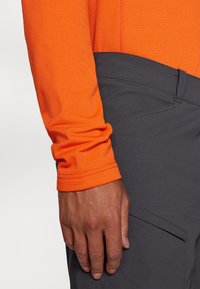 Ilgi piedurkņu oranž tops ar teksturētu audumu, apvienots ar tumši pelēkām biksēm, kam ir elegantais dizains un sānu kabatas detalizācija.