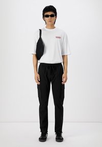 Bílé tričko s červeným textem, černé cargo kalhoty s bočními kapsami a černé tenisky. Doplňky zahrnují černou crossbody tašku a sluneční brýle.