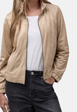 Frau trägt beige Wildleder-Reißverschlussjacke über weißem Hemd und schwarzen High-Waist-Jeans, eine Hand in der Tasche, vor einfachem Hintergrund.
