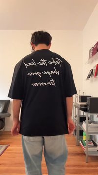 Mees seisab toas kaamera poole seljaga, seljas must suur T-särk valge kaldkirjas tekstiga ja heledad sinised teksad elutoas.