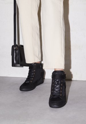 Person trägt cremefarbene Hose, schwarze Keilsneaker mit Schnürsenkeln und Riemen und hält eine schwarze Lederhandtasche an der Seite.