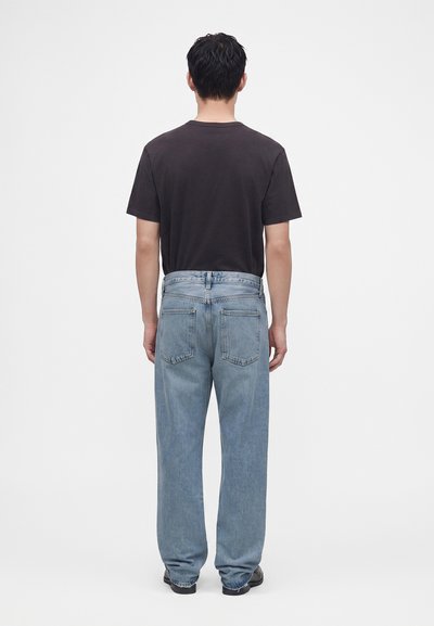 Homme portant un t-shirt noir et un jean large bleu clair, debout, de dos, contre un fond uni clair.